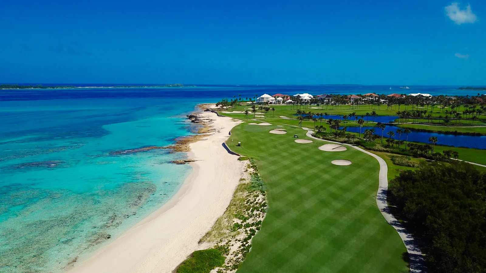 From above, the green fairways of a golf course running beside a strip of white sand and turquoise sea.
