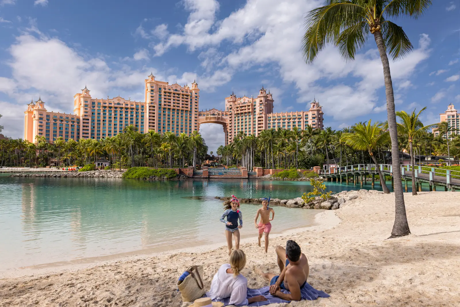 A family on the sand of a lagoon beneath a palm; on the far shore, The Royal’s towers with the bridge between its 2 wings.
