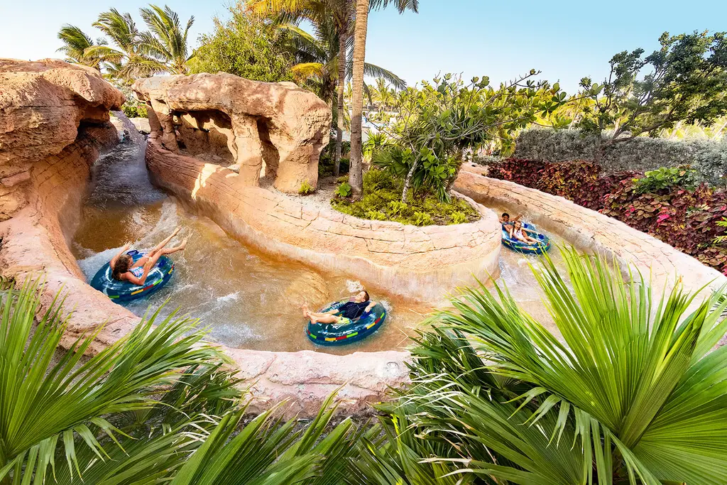 A man-made river between rocks and palms, with 3 people on inflatable tubes carried by the current.