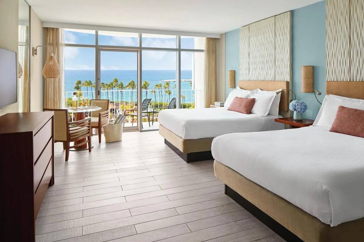 A Premium room at The Coral with 2 queen beds, a pale blue wall and a glazed door to a balcony with a sea view.