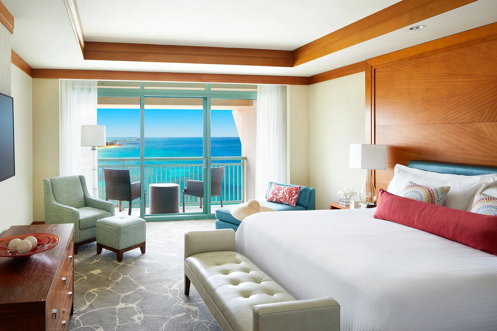 The bedroom of an Azure suite, with a king bed, a pale leather bench and a furnished balcony over turquoise sea.