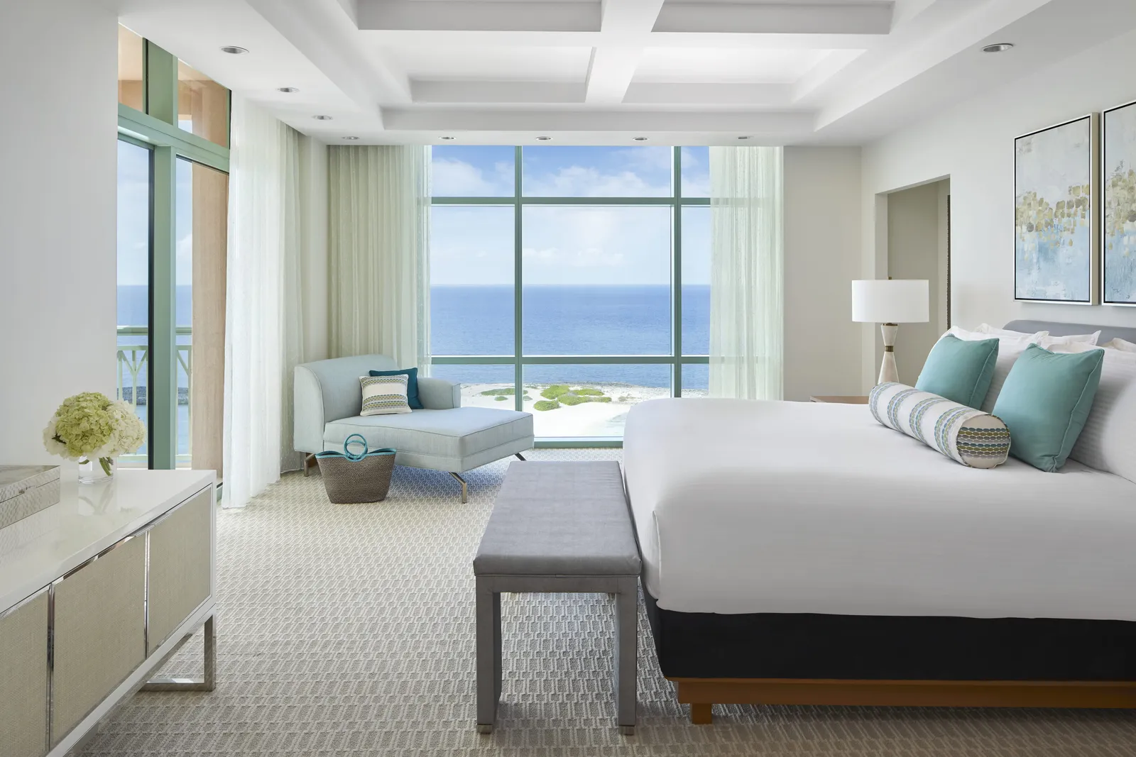 The bedroom of a Sapphire suite, in white and pale blue, with a chaise longue by a corner window over the sea.