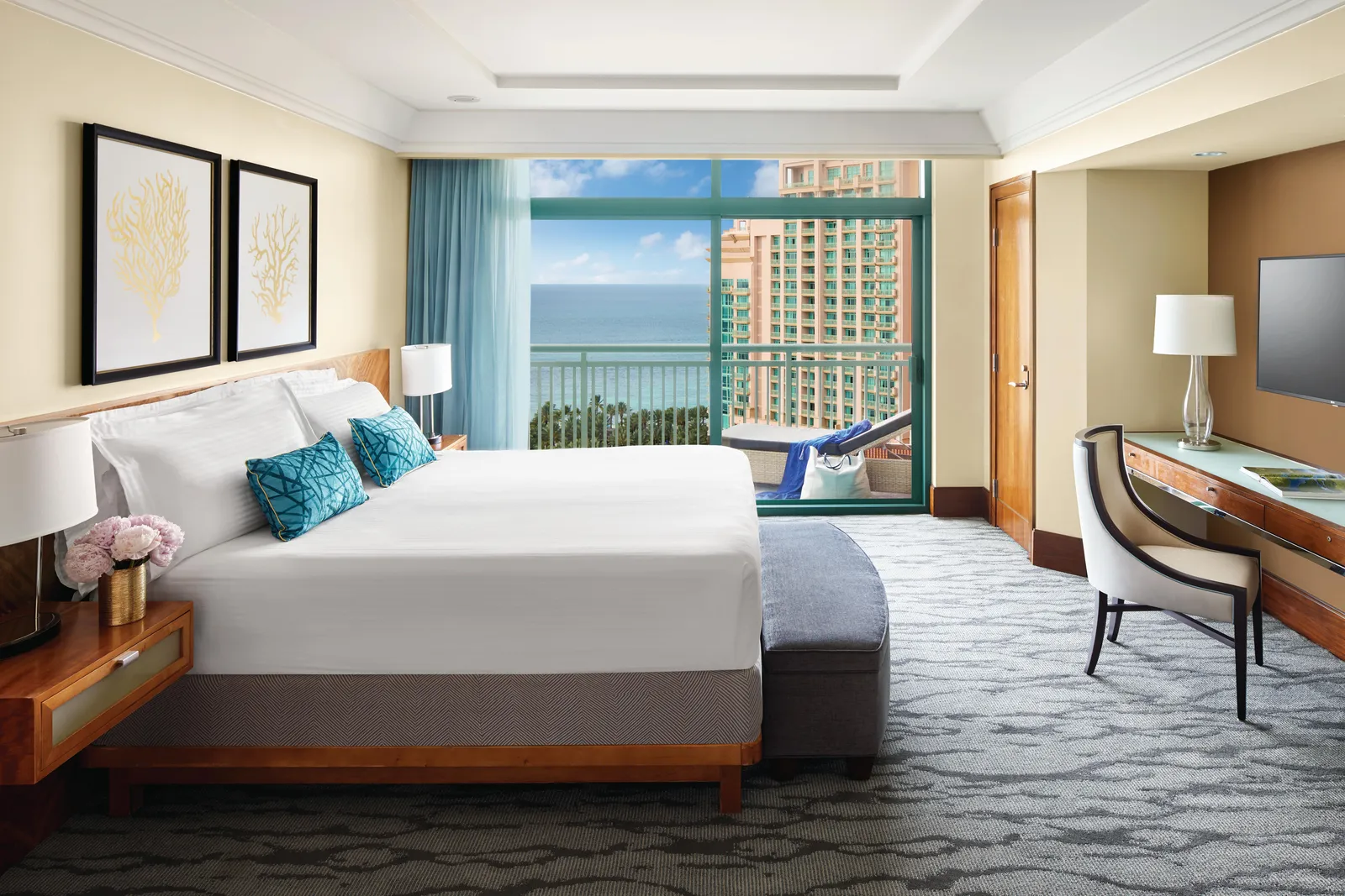 The bedroom of a one-bedroom suite at The Reef, with a king bed, a desk and a window onto the sea and The Cove’s tower.