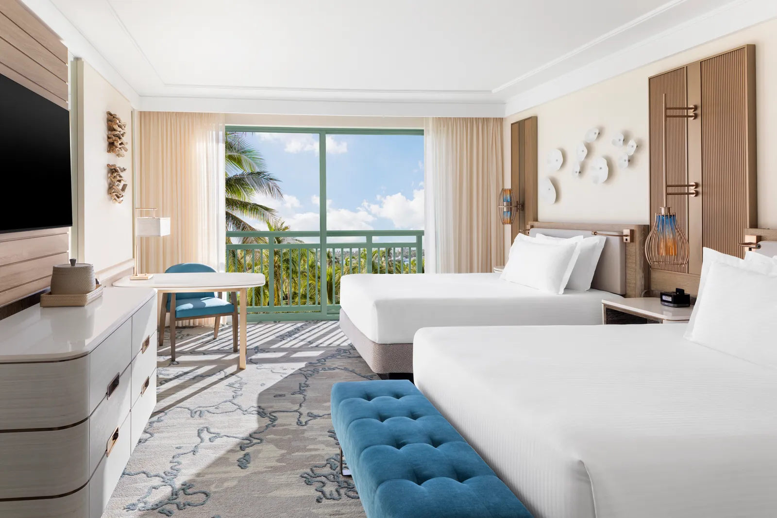 A room at The Royal with 2 queen beds, a blue bench at their foot and a full-height window onto palms.
