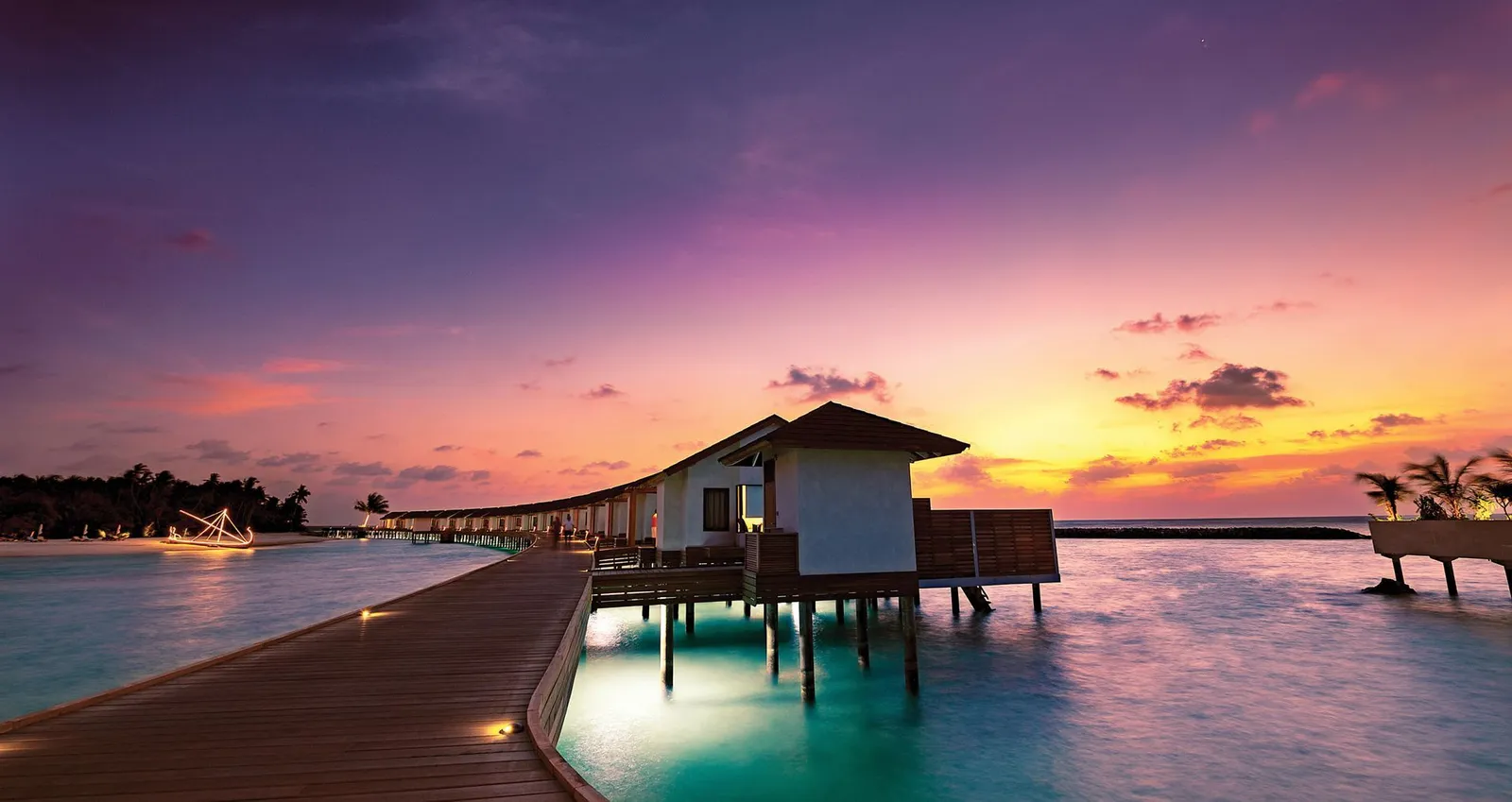 A Kanifushi Water Villa at dusk, its lit walkway above a pink lagoon