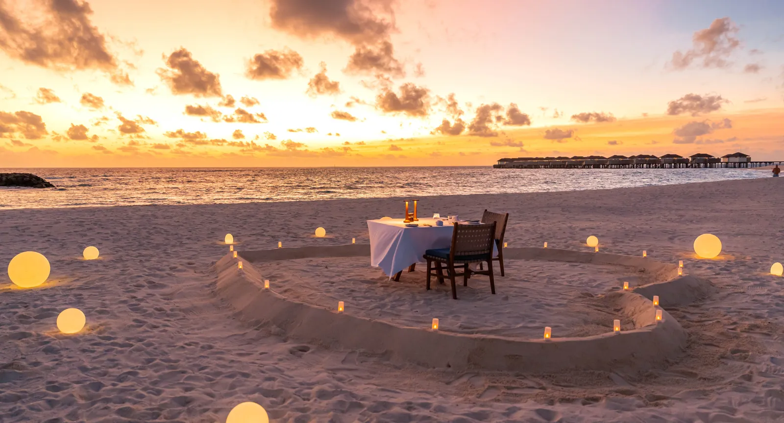 A private dinner on the beach at sunset, inside a heart traced in lanterns.