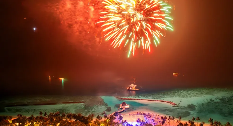 Fireworks at night over the lagoon, with the lit beach and villas below.