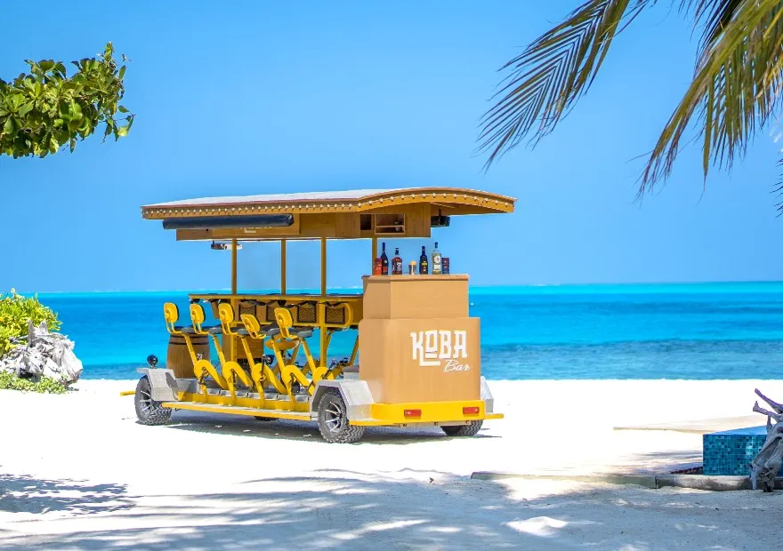 KOBA, the pedal-powered bar, parked on the white beach before the turquoise sea.