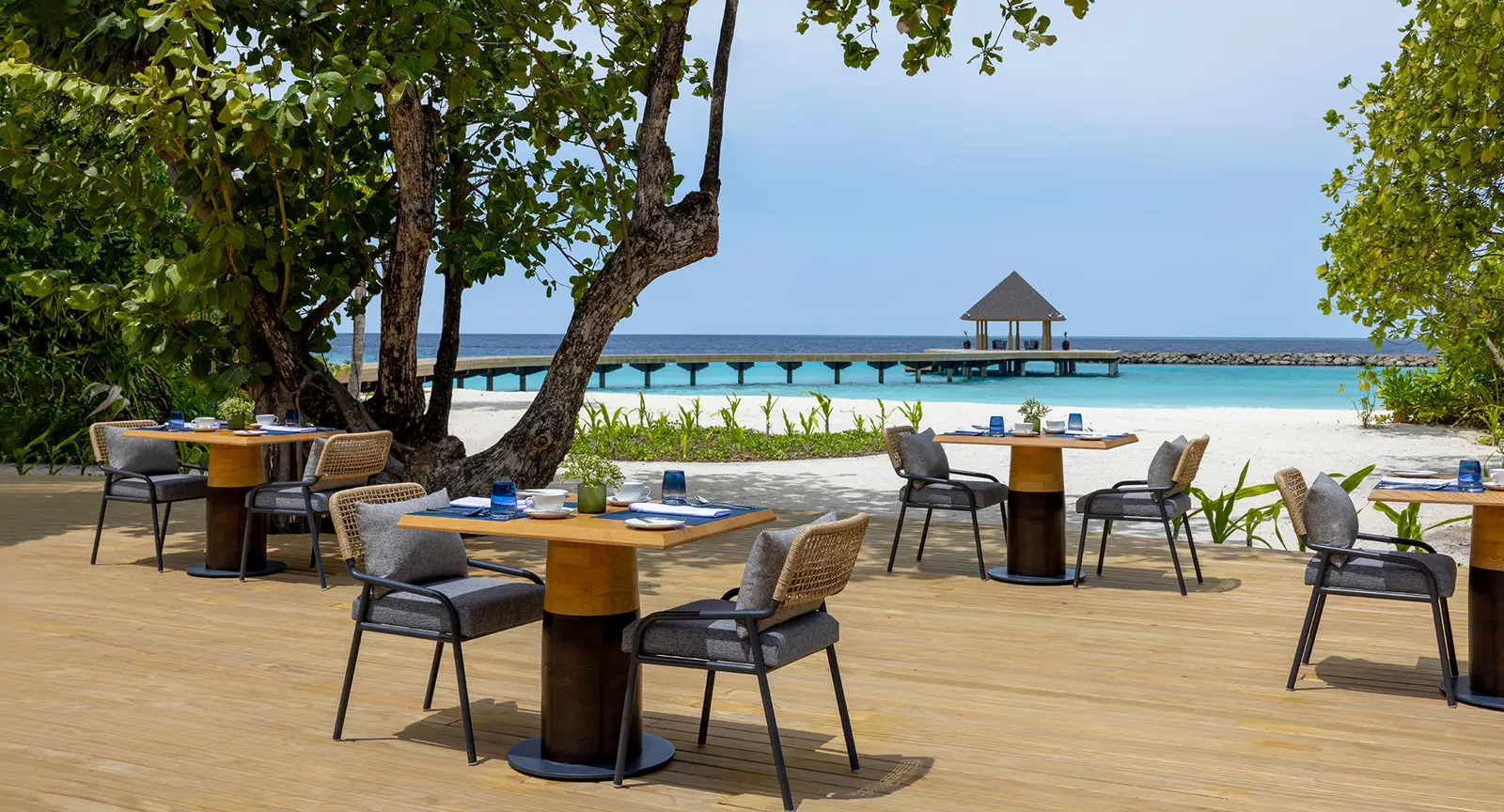 The Ocean Terrace deck on the beach, tables in the shade of the trees and the jetty beyond.