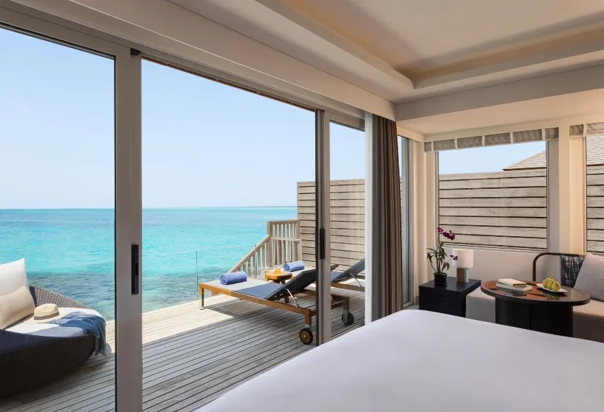 An Over Water Villa bedroom: the bed, the daybed on the deck and the lagoon beyond the glass.