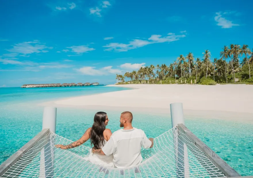 A couple sitting on the net slung over the turquoise water, with the beach and palms ahead.