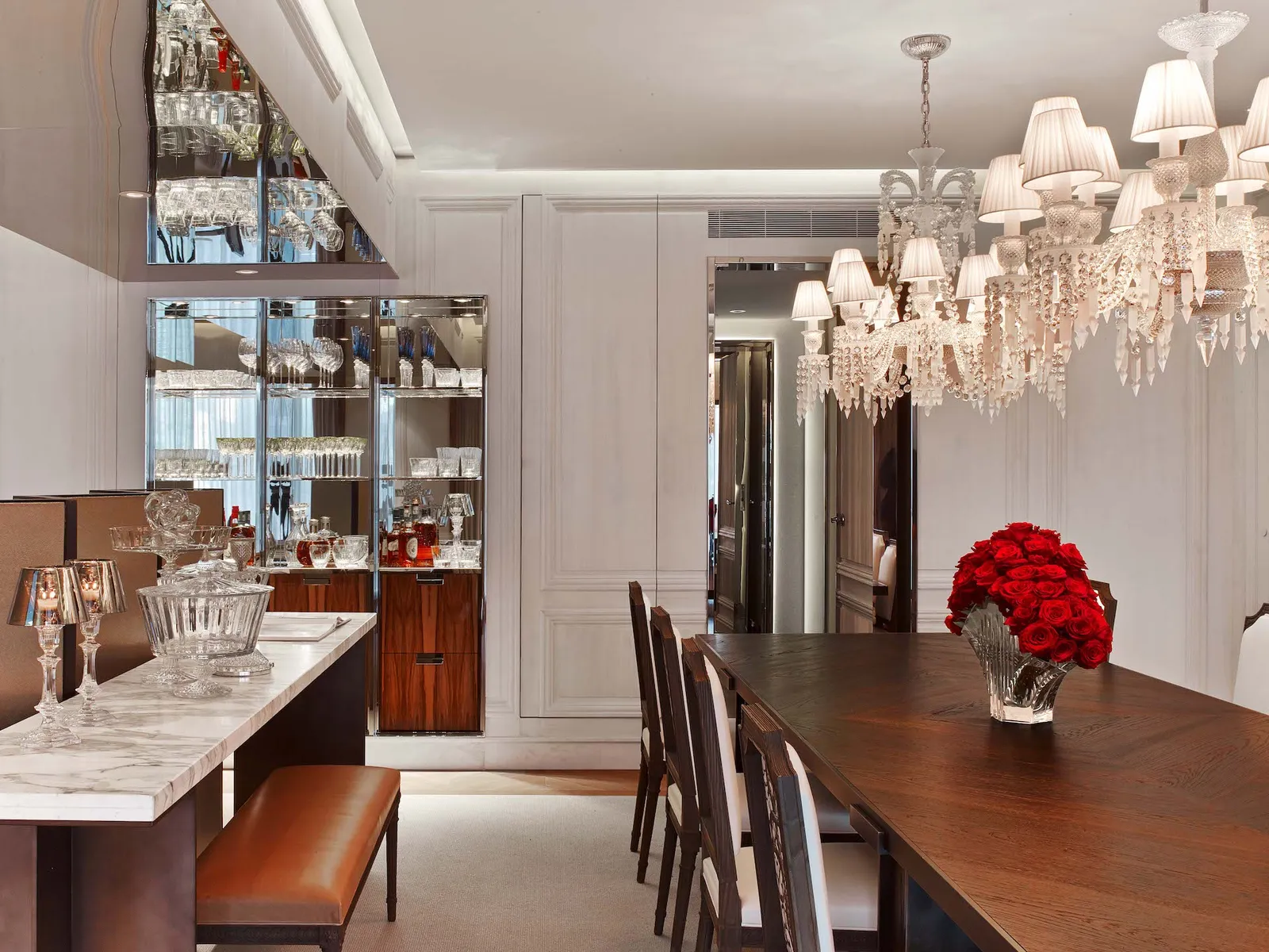 A dining table beneath a crystal chandelier, with a living room beyond
