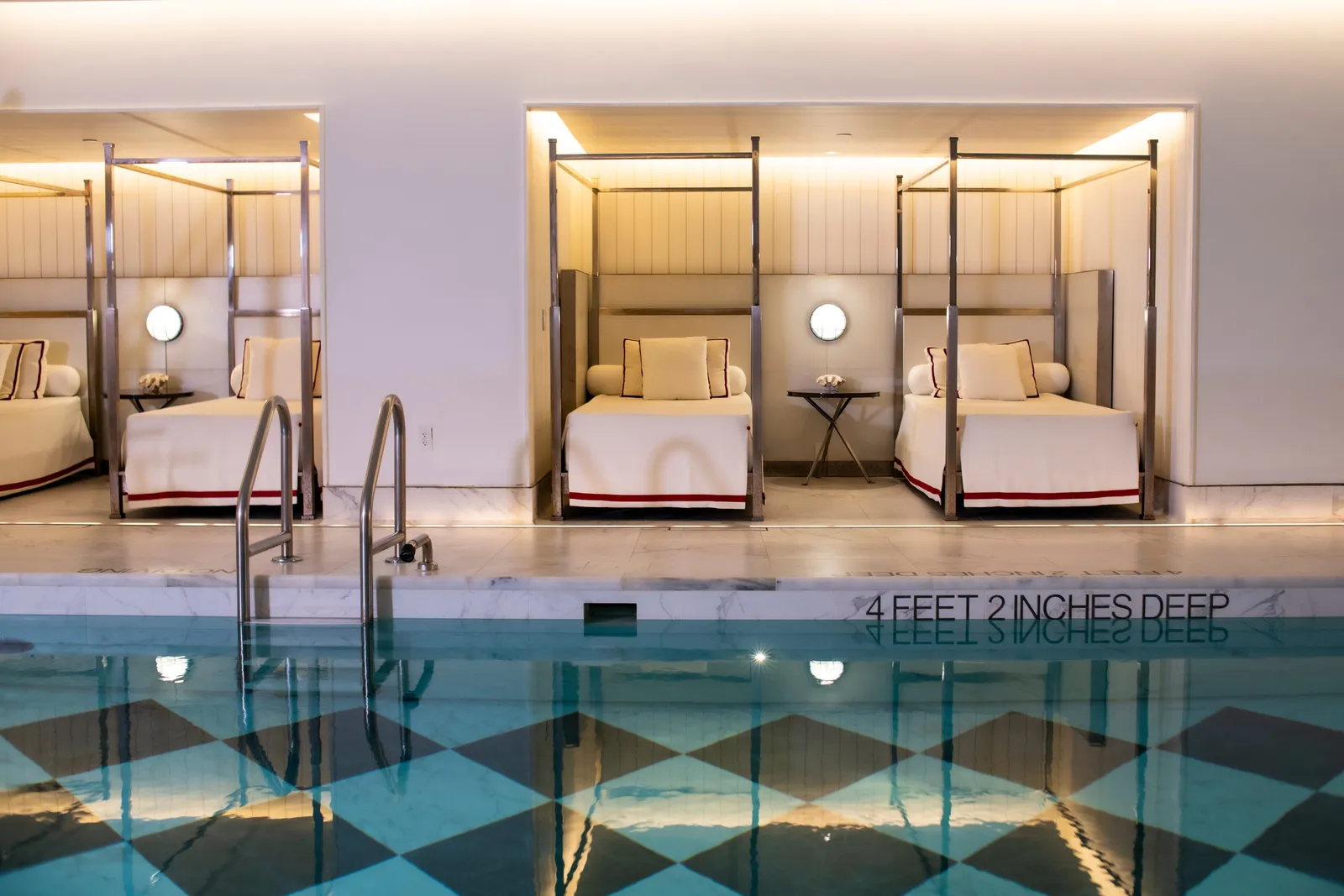 Indoor pool with checkerboard tile flooring, poolside cabana daybeds, and depth marker reading 4 feet 2 inches.