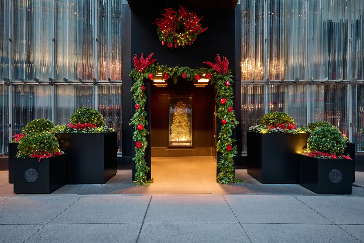 Baccarat Hotel entrance decorated with holiday garland, red wreath, and topiaries, framing illuminated gold tree inside