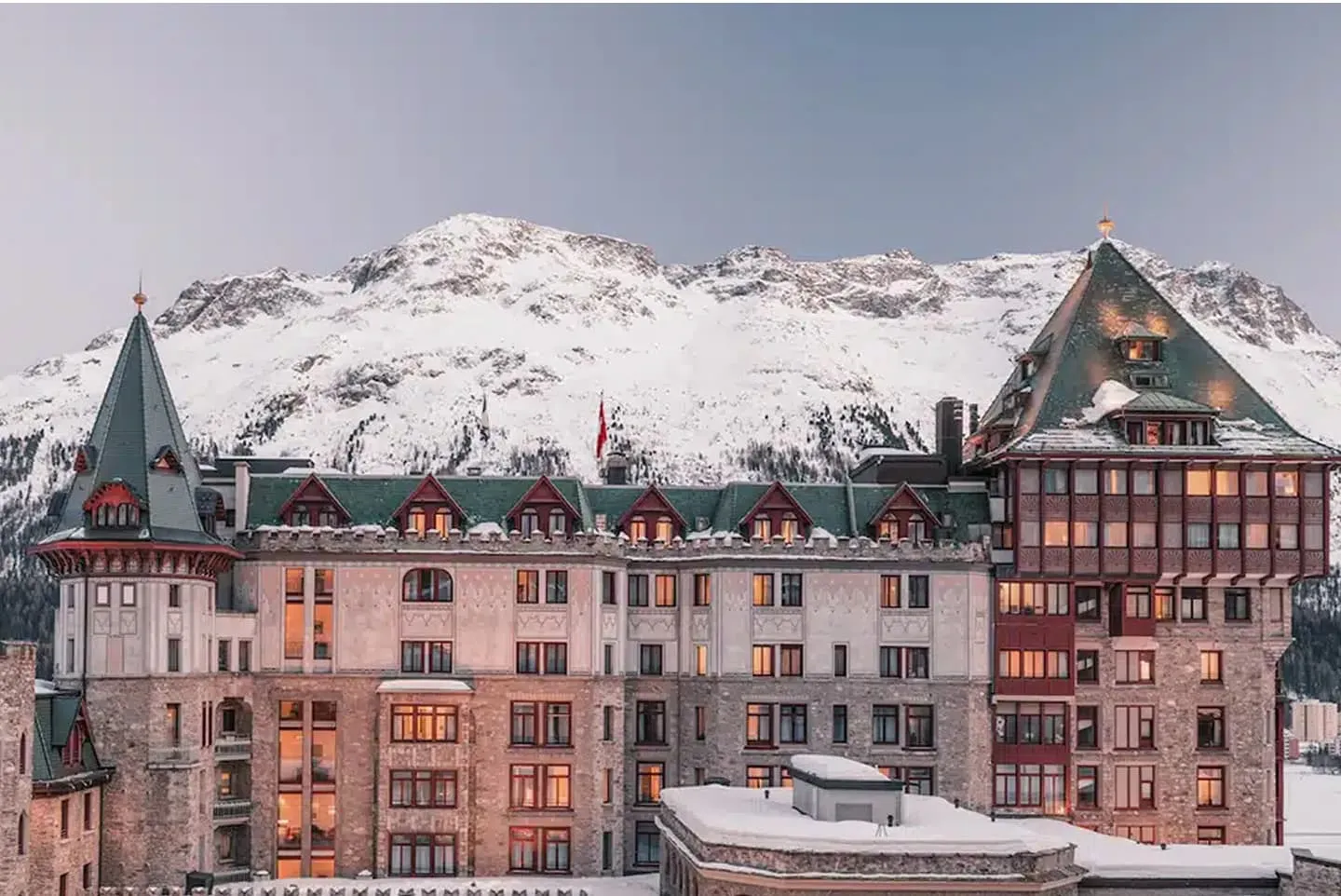 The façade of Badrutt’s Palace in winter, with its windows lit, pointed green roofs and a snow-covered mountain behind.