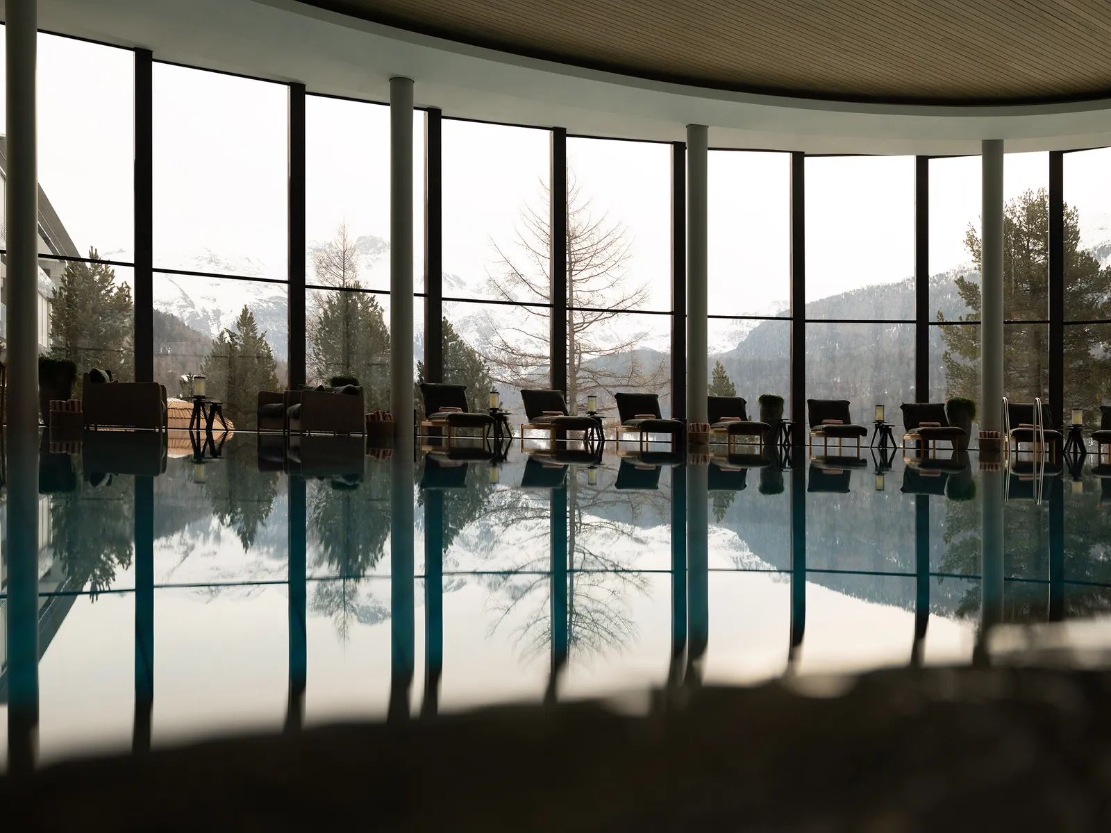 The indoor pool with its full-height windows, loungers along the edge and the larches and mountains reflected in the water.