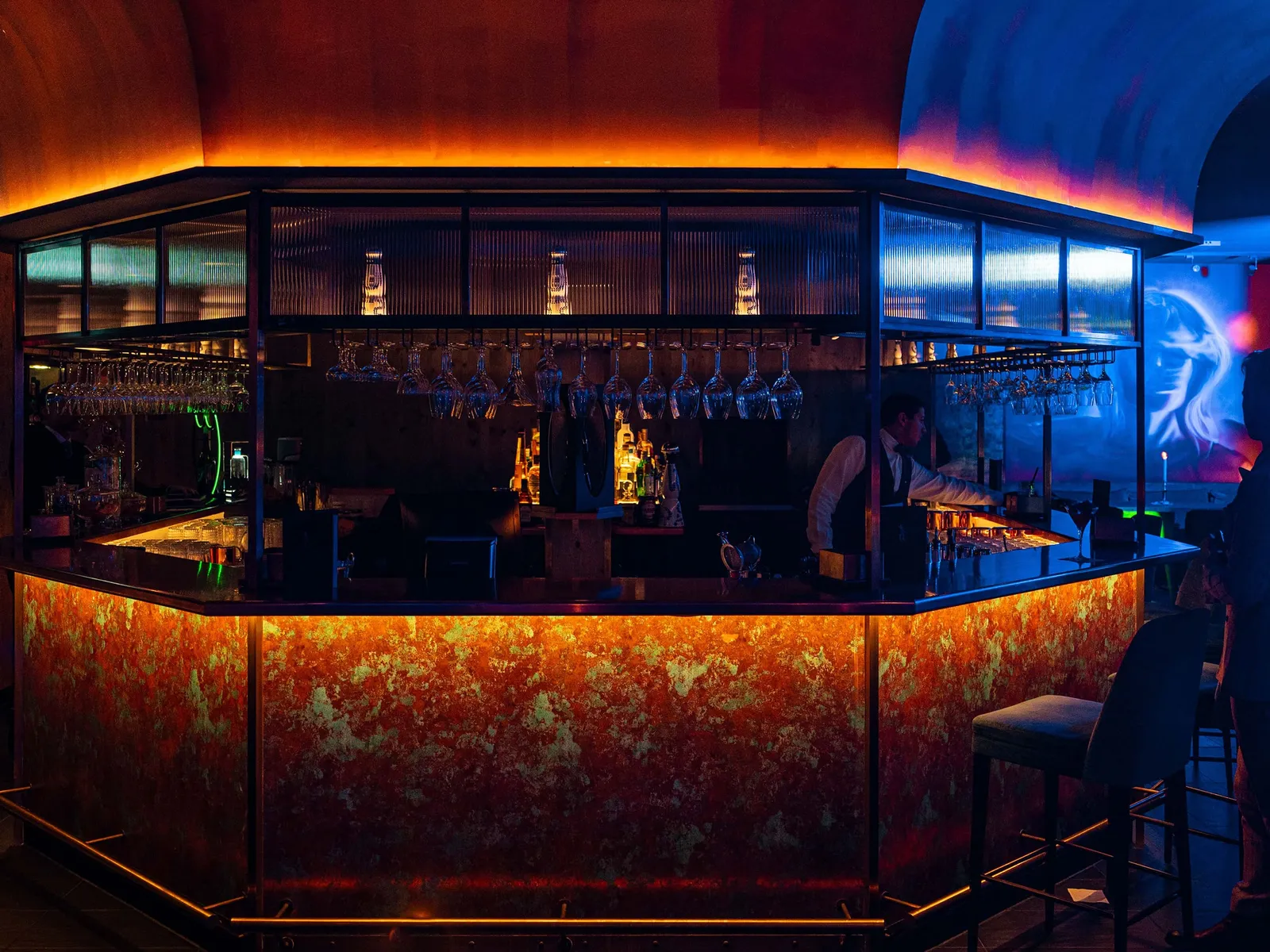 The bar at King’s Social House, lit in orange and blue, with bottles in the cabinet and a bartender at work.
