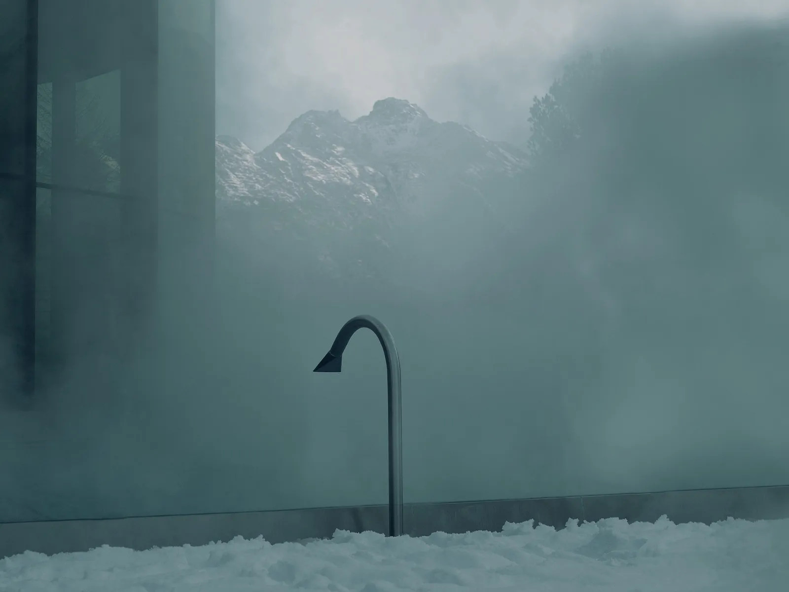 The outdoor pool in winter: steam above the warm water, a swan-neck jet, snow on the edge and mountains in the haze.