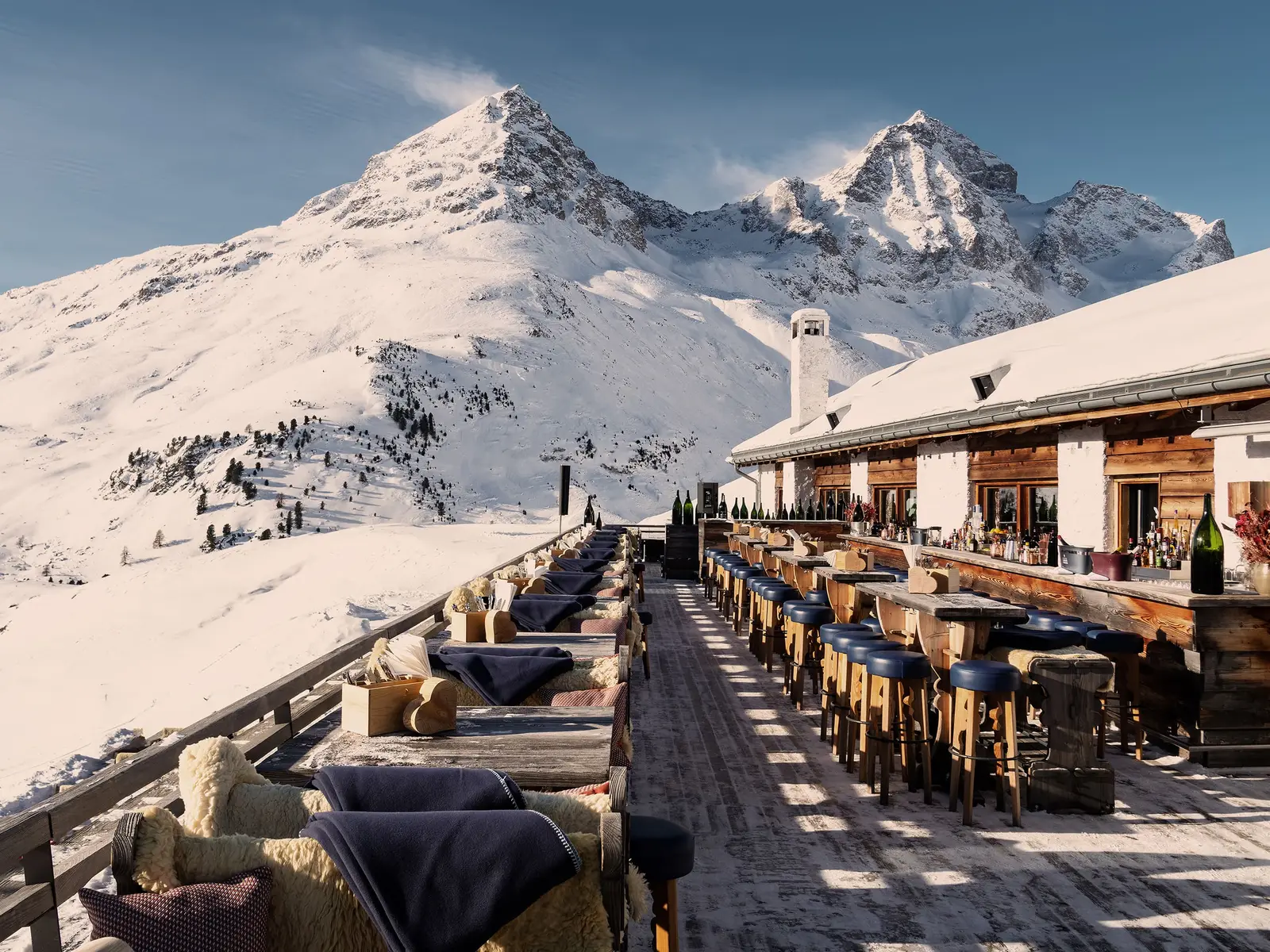 The terrace at Paradiso on Corviglia: tables and sofas with blue blankets in a row on the decking, the chalet and the snowy peaks.