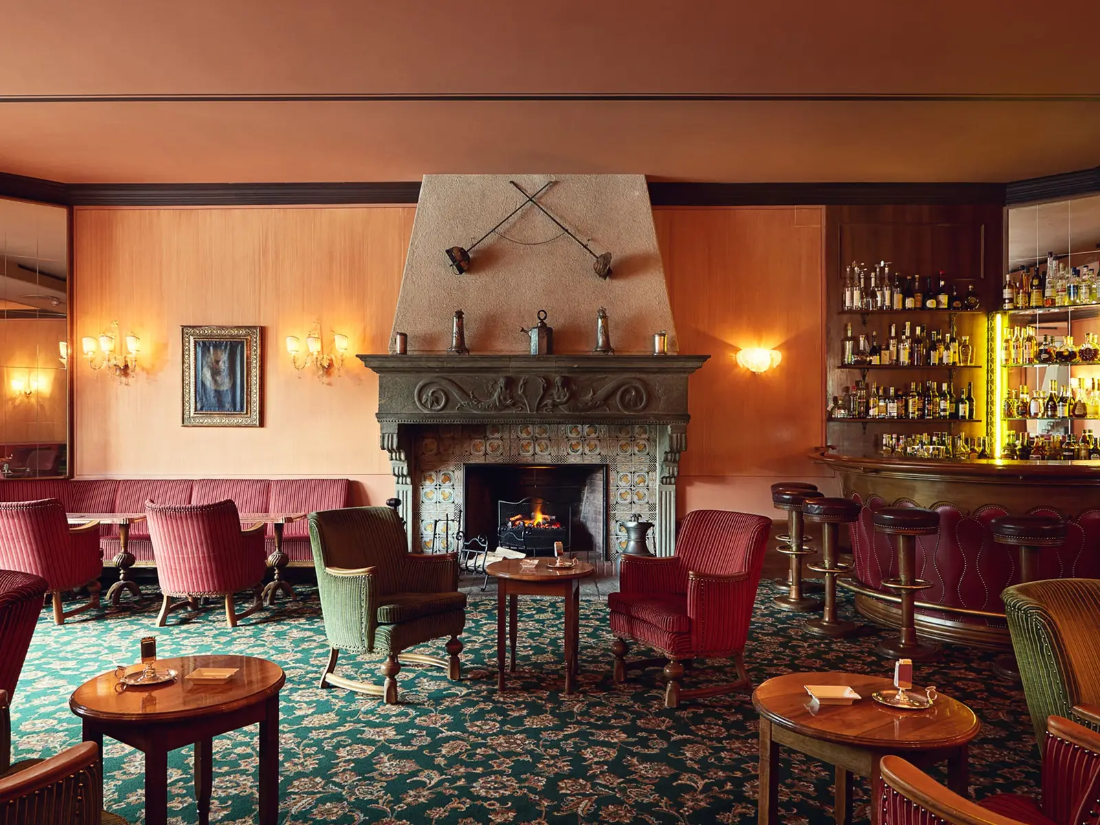The Renaissance Bar: the fire lit, red velvet armchairs on the green carpet and the bar with its bottles on the right.