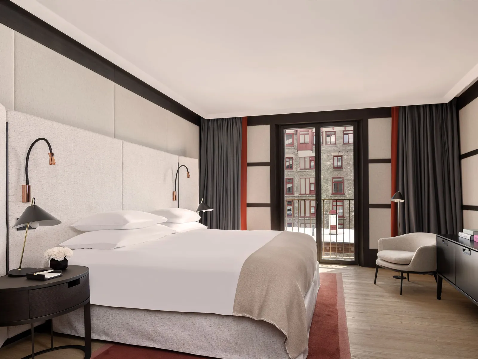 A Serlas Grand Room: the bed with its fabric headboard, black reading lamps, an armchair and the French window onto the village houses.