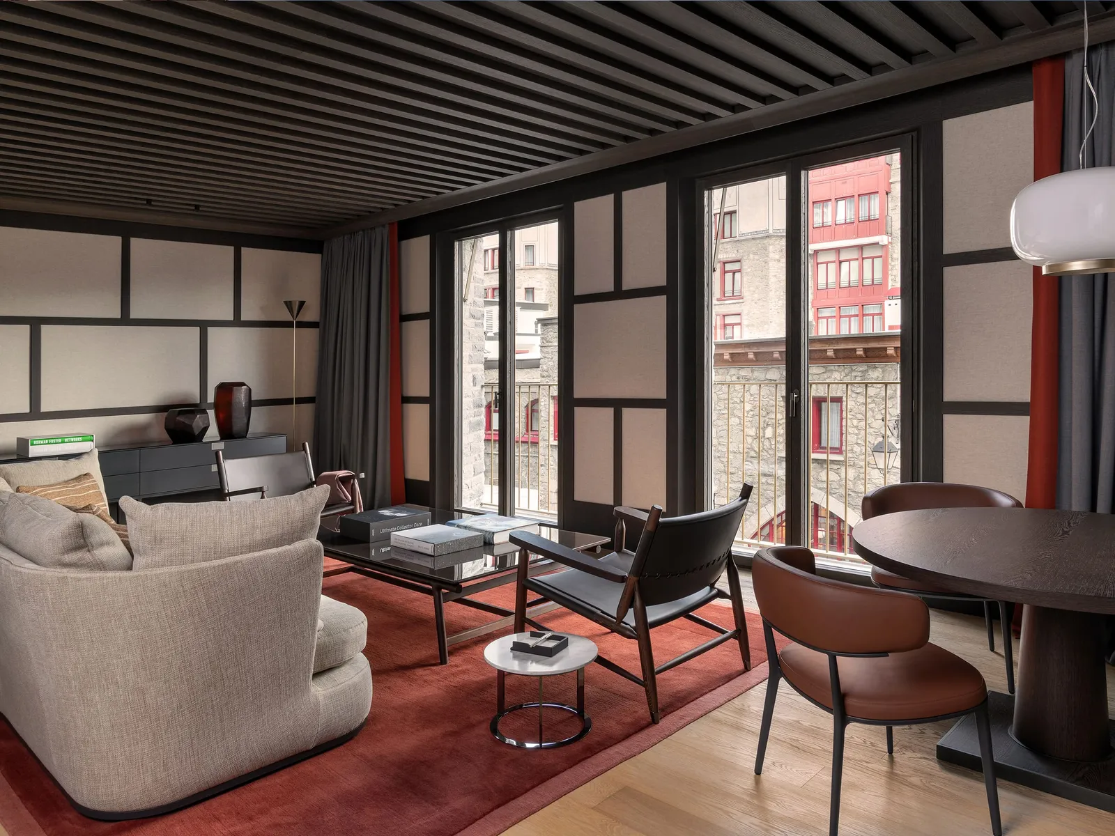 The living room of a Serlas Suite: the dark beamed ceiling, a round table with leather chairs, the red rug and windows onto the houses opposite.