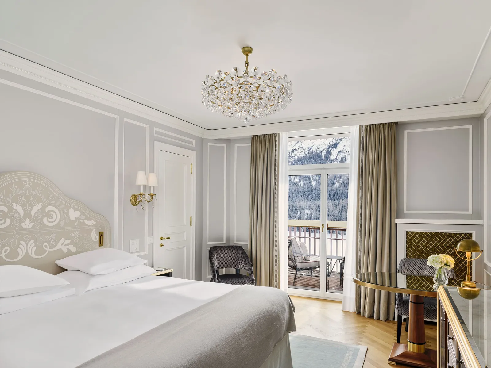 A Signature Double Room: the bed with its carved headboard, the crystal chandelier and the French window to the balcony and the snowy mountains.