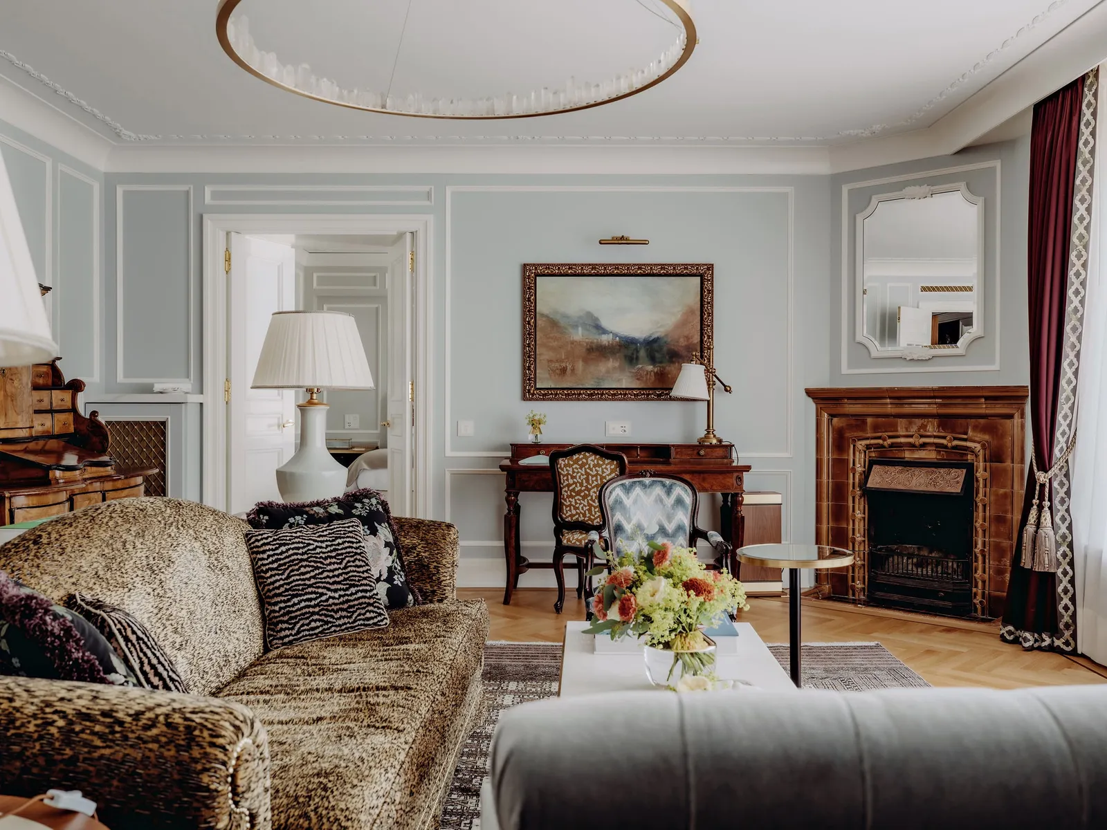 The living room of a St. Moritz Suite: the leopard-print sofa, the tiled fireplace, a writing desk and a ring-shaped ceiling light.