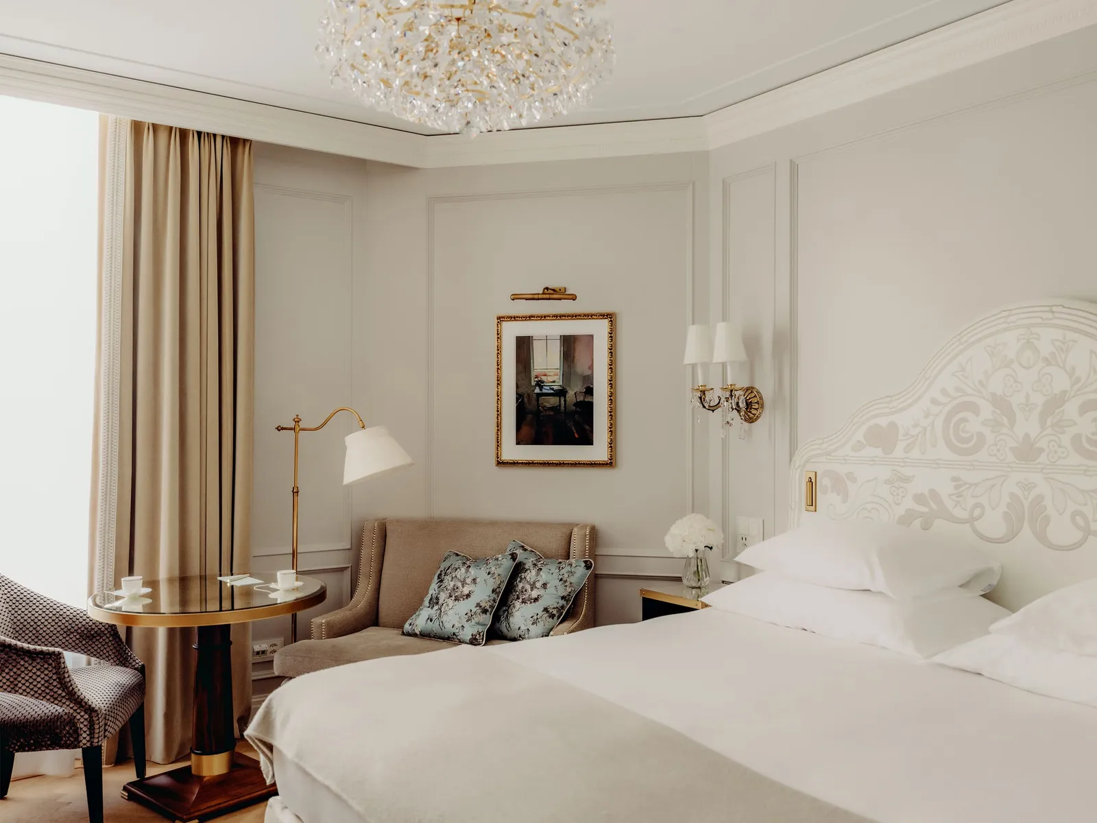 A Superior Double Room: the bed with its white fretwork headboard, an armchair, a round table and the crystal chandelier.