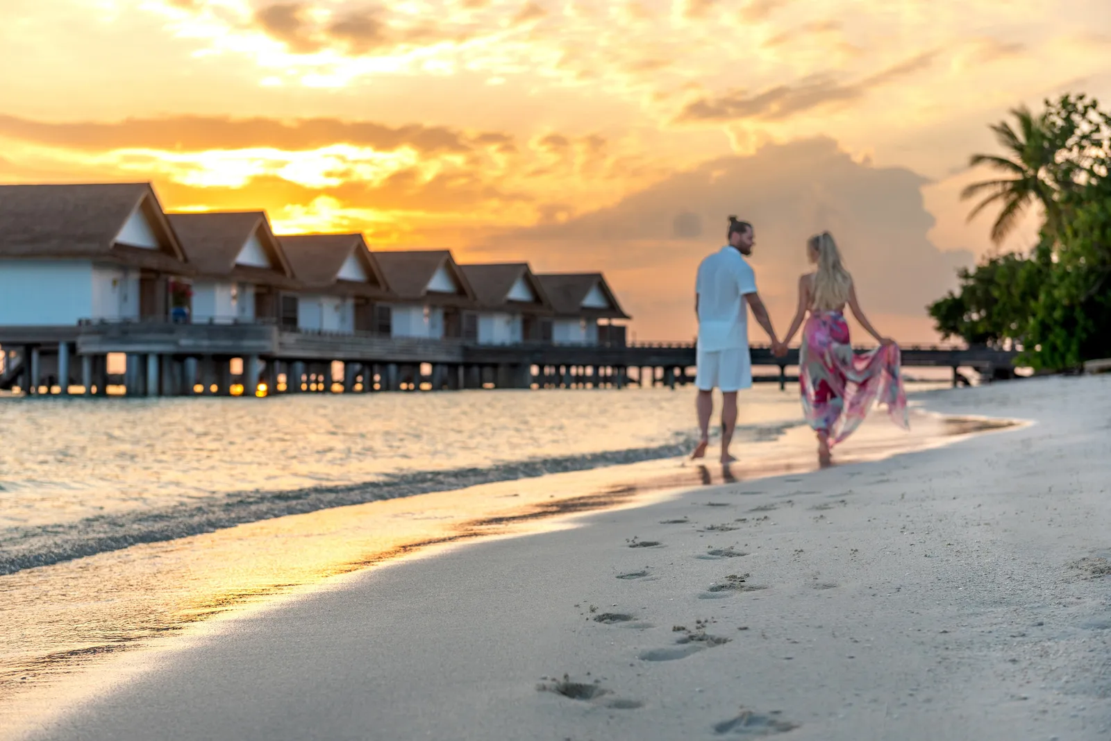 A couple walking along the beach at sunset, the row of overwater villas behind them