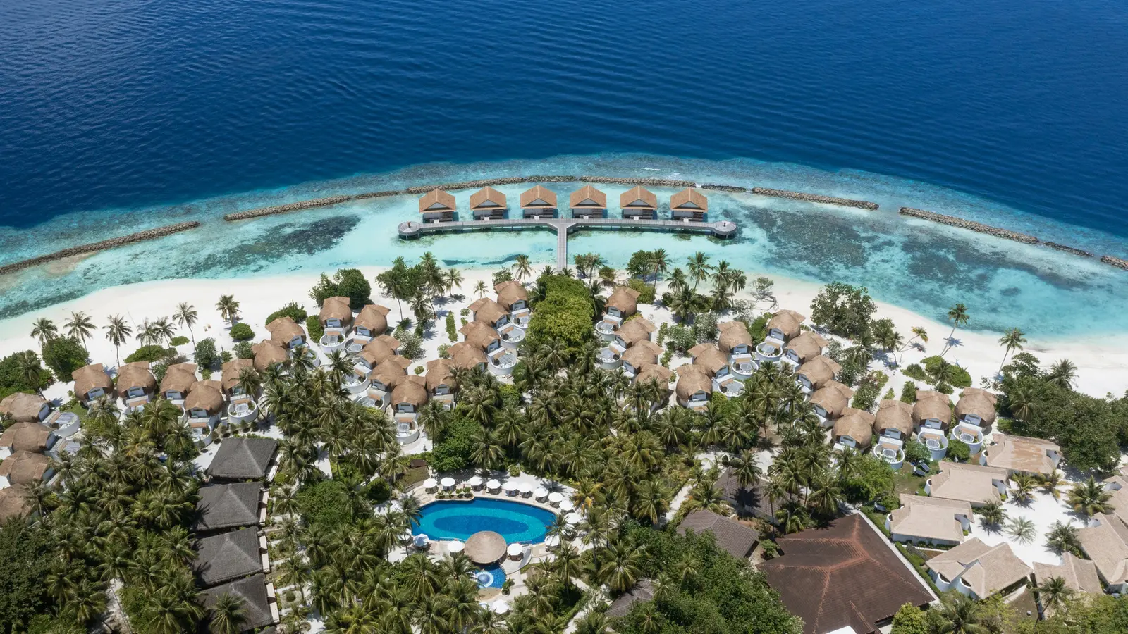 The resort from the air, villa roofs among the trees, the pool and the overwater villas beyond the beach