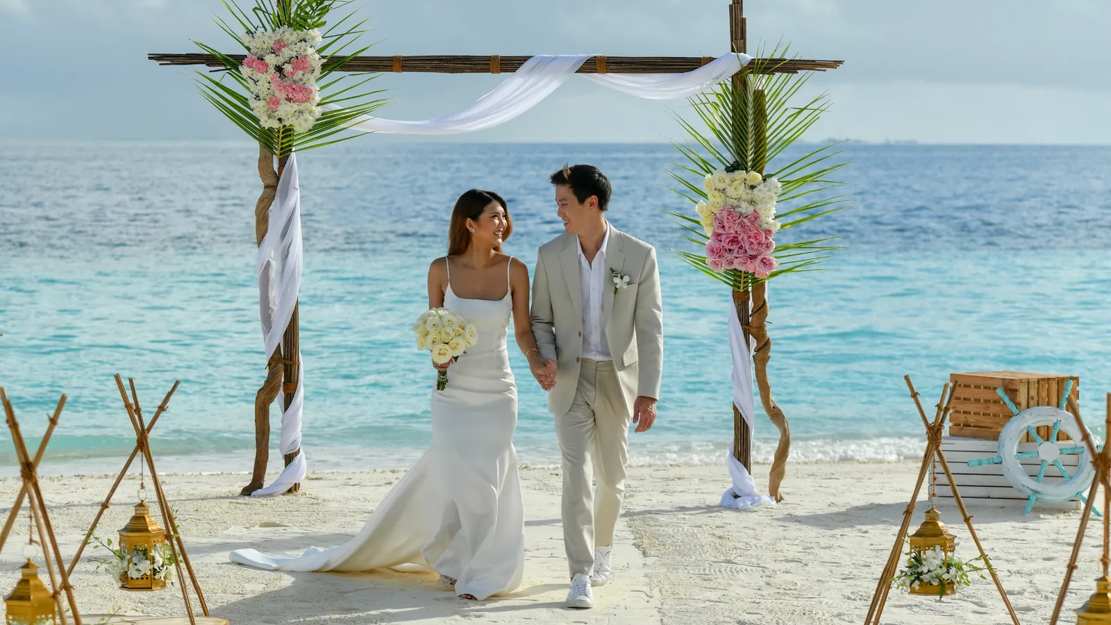 A newly married couple walk on the sand after the ceremony, under the floral arch, the sea behind them.