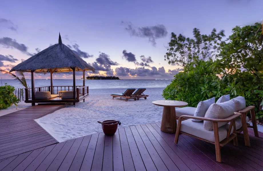 A Beachfront Sunset View Pool Villa’s deck at dusk, with its thatched sala and loungers on the sand facing the sunset.