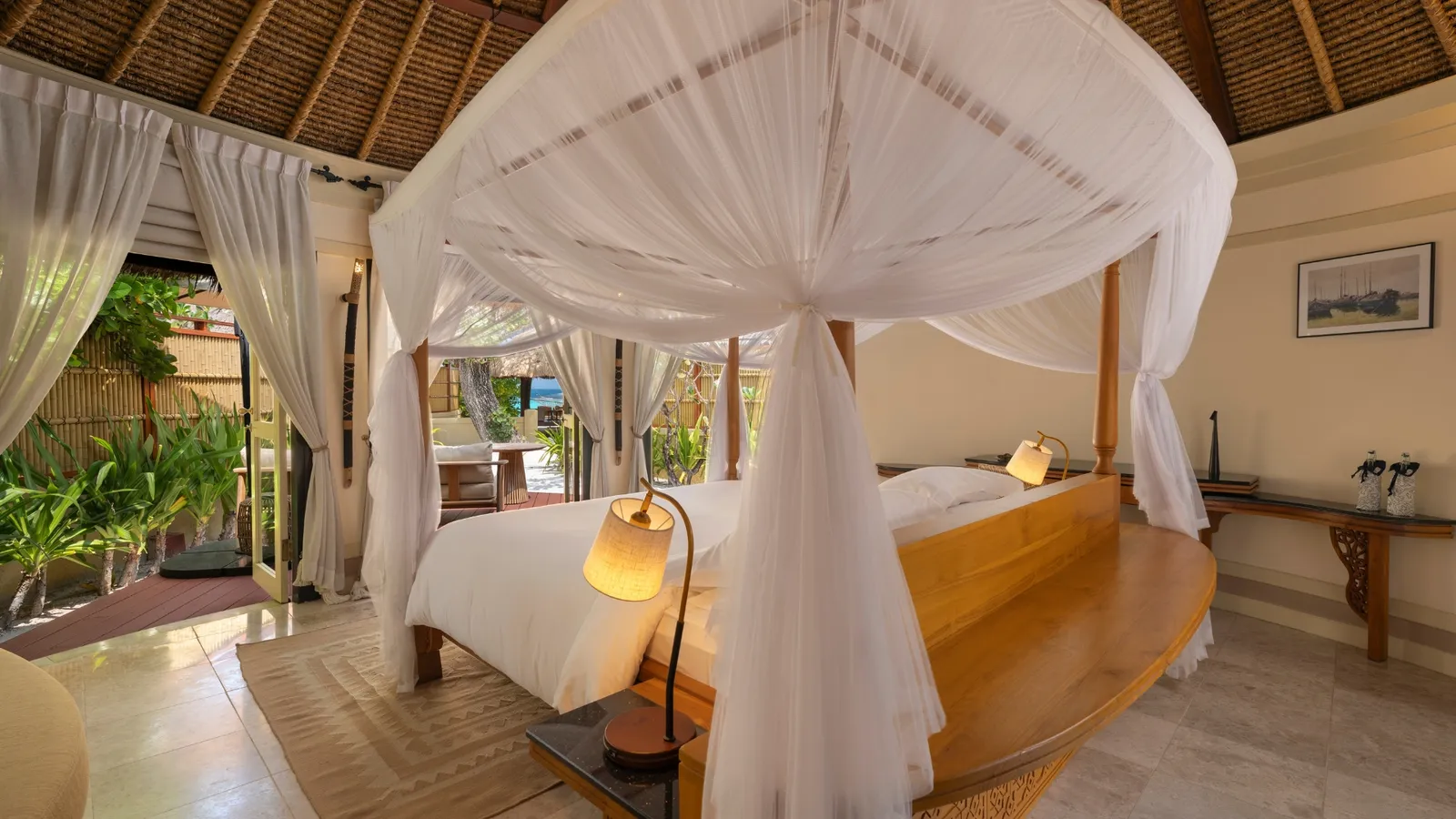 An Oceanview Pool Villa bedroom: the four-poster bed with white drapes, the timber bath and the deck beyond the open doors.