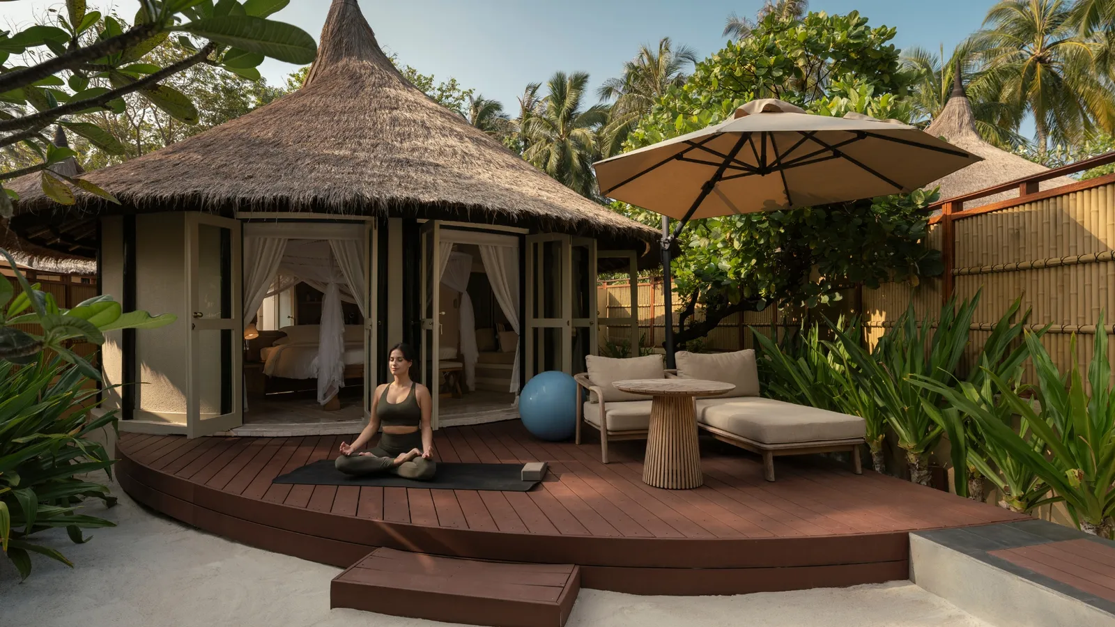 A Wellbeing Sanctuary Pool Villa: the deck among the greenery, a guest meditating on the timber and the thatched villa behind.