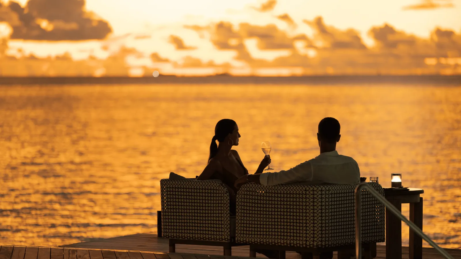 A couple on the sofa of a beach sala, in silhouette against an orange sunset over the lagoon.