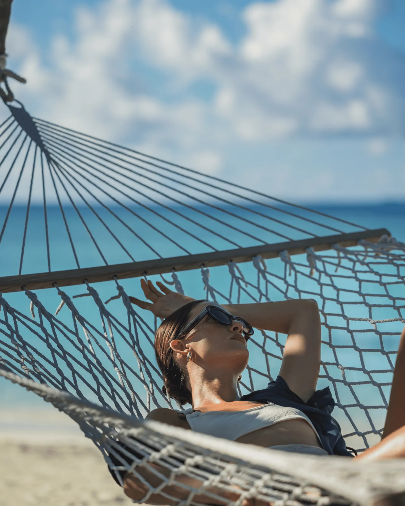 A guest lying in a rope hammock facing the sea, under a blue sky.