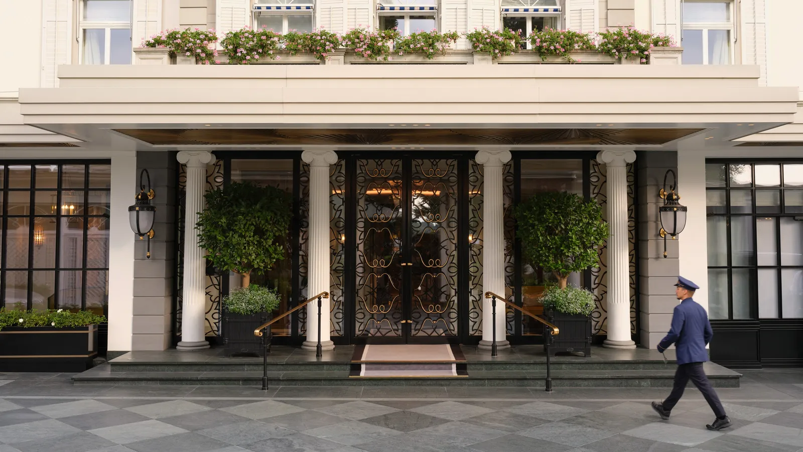 The hotel’s entrance on Talstrasse: a portico with columns and potted trees under balconies of flowers, and a doorman in livery walking past
