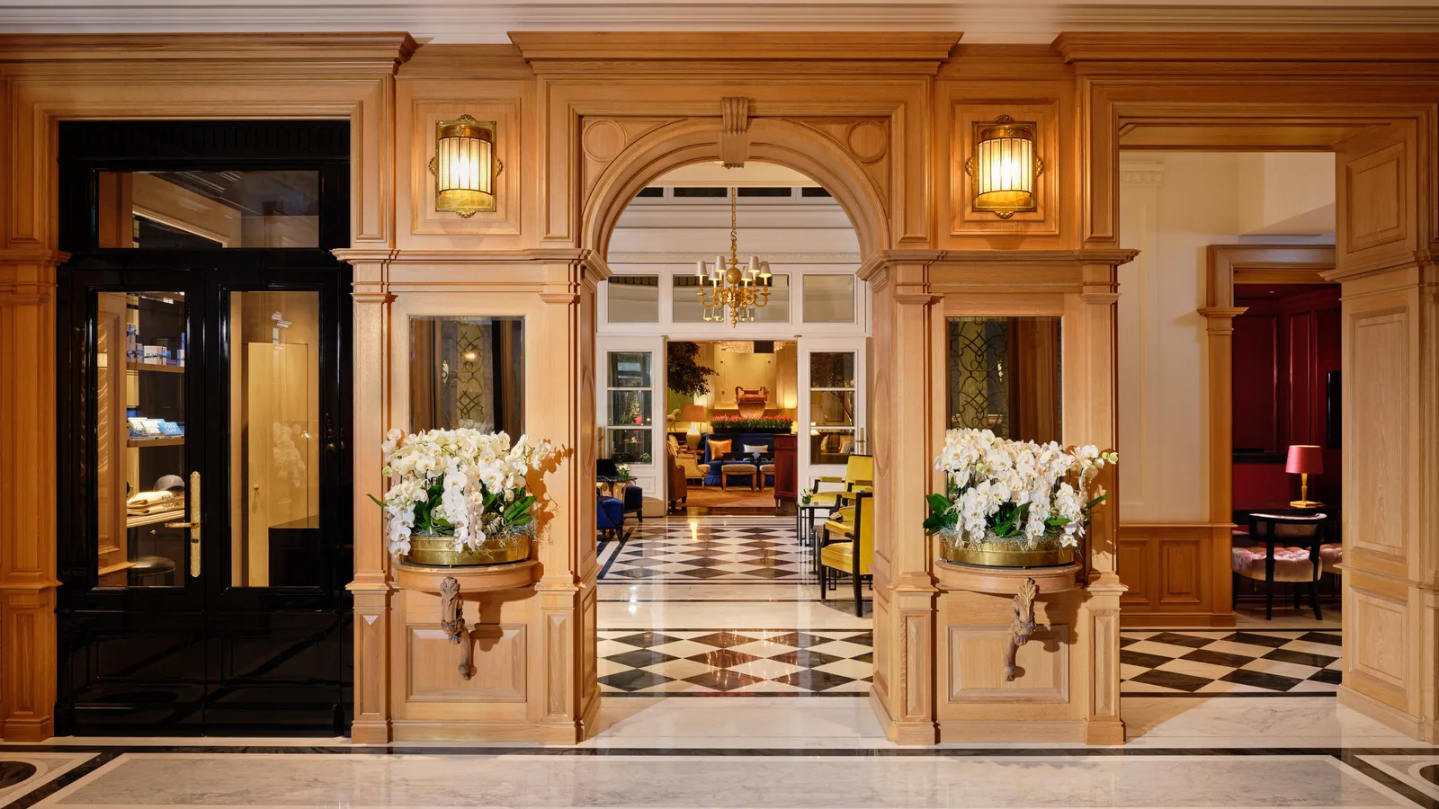 The lobby seen through the entrance arch: carved wood panelling, white orchids, a black and white marble floor and a chandelier beyond