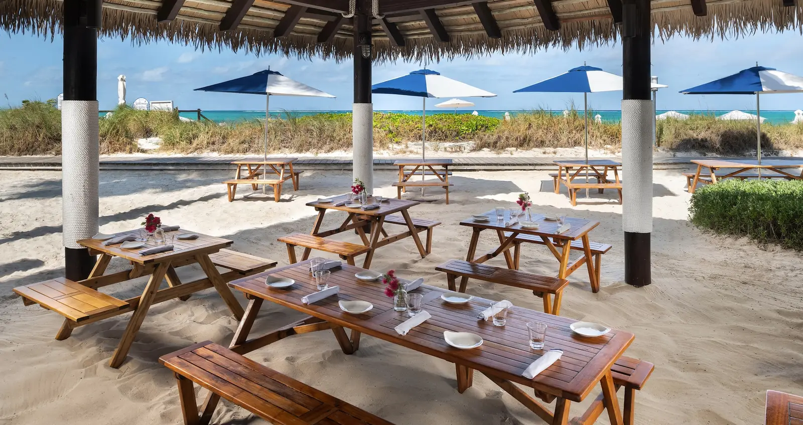 Wooden tables and benches laid for a meal on the sand, under a thatched roof, with blue umbrellas and the sea beyond.