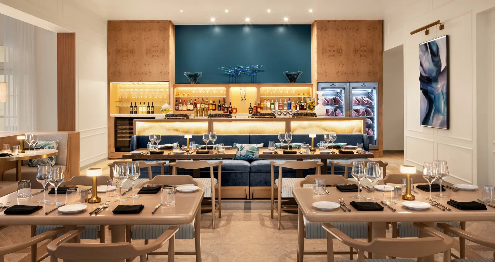 A dining room with pale wooden tables laid for dinner, a bar with a dark blue back wall and 2 glass-fronted cabinets for ageing meat.