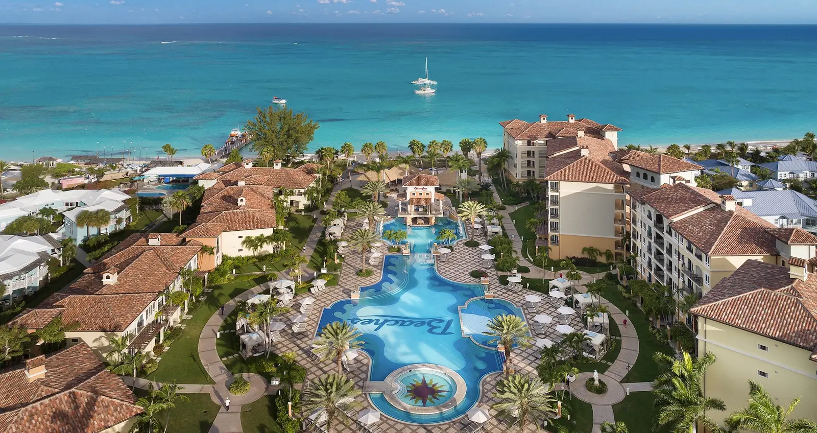 From the air, the terracotta-roofed buildings of the Italian Village around a large curving pool among palms, with the beach and the turquoise sea just behind.