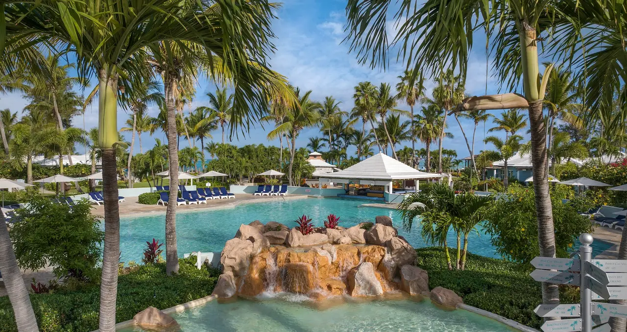 A pool among palms with a rock island and a small waterfall at its centre, blue loungers along the edge and a white pavilion.