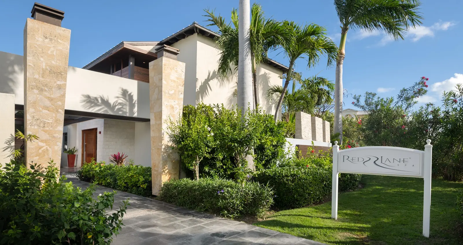 The entrance to Red Lane Spa: a path between lawn and palms leads to a low white building with a stone wall and the spa’s sign.