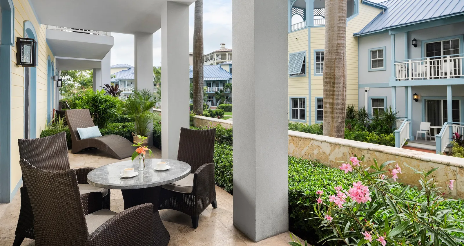 A ground-floor veranda with woven armchairs, a round table and 2 loungers, facing the garden and the pastel houses.