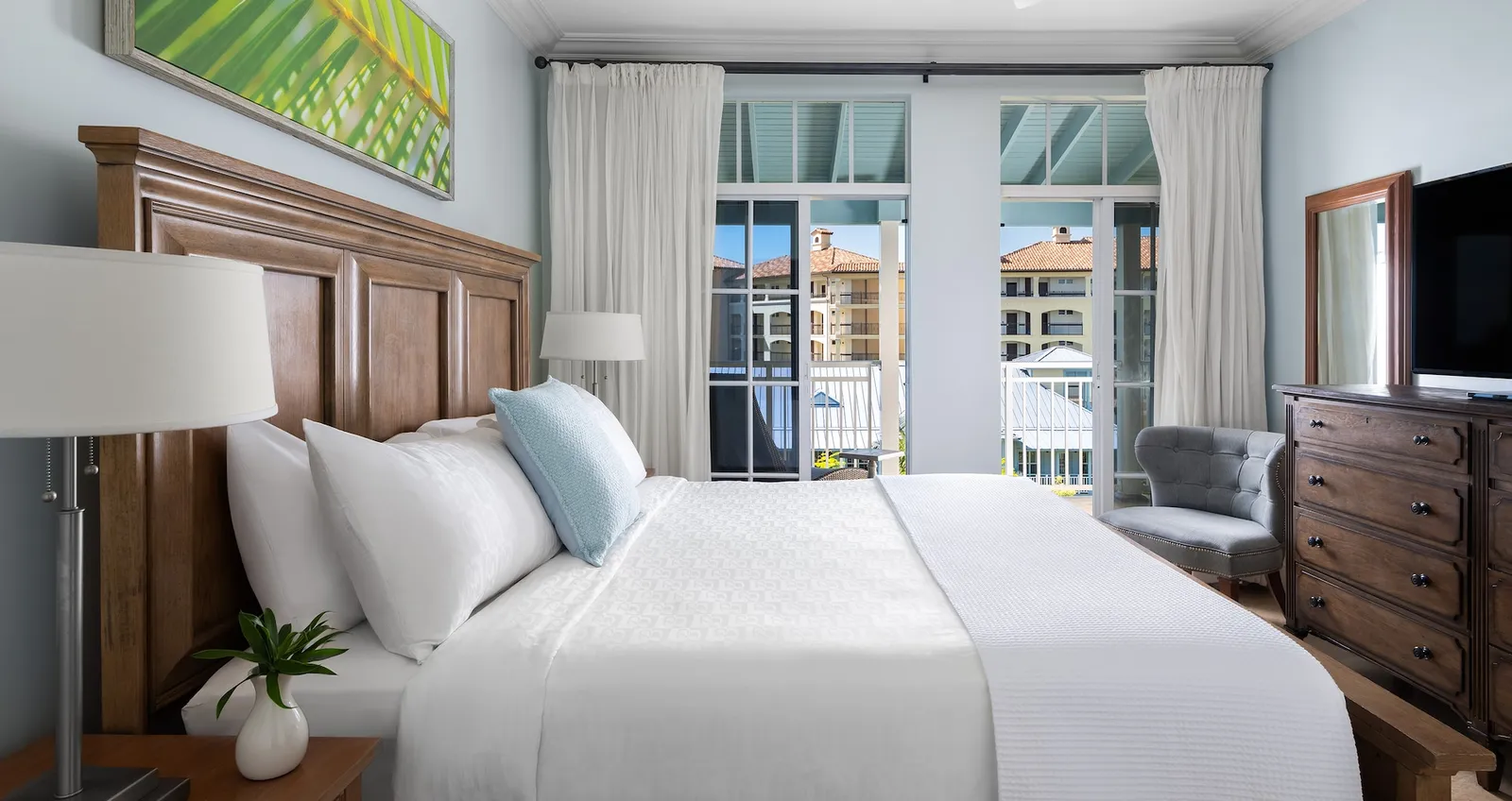A bedroom with a king bed, a wooden headboard, the chest of drawers with the television and the doors to the balcony.