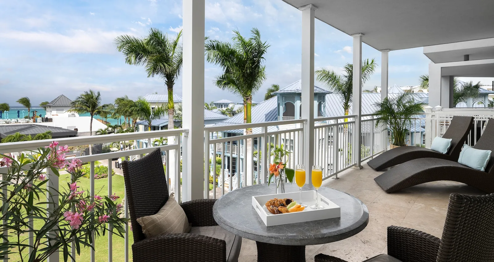 A balcony with a round stone table, woven armchairs and 2 curved loungers, above roofs, the pool and, further off, the sea.