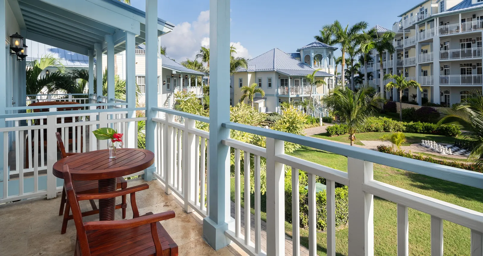 A balcony with a red wooden table and chairs and a white railing, above lawns and white-and-blue buildings.