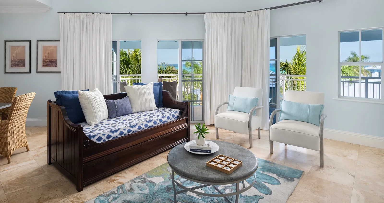 A bright living room with a dark wooden daybed, 2 white armchairs, a round coffee table and tall windows with white curtains.