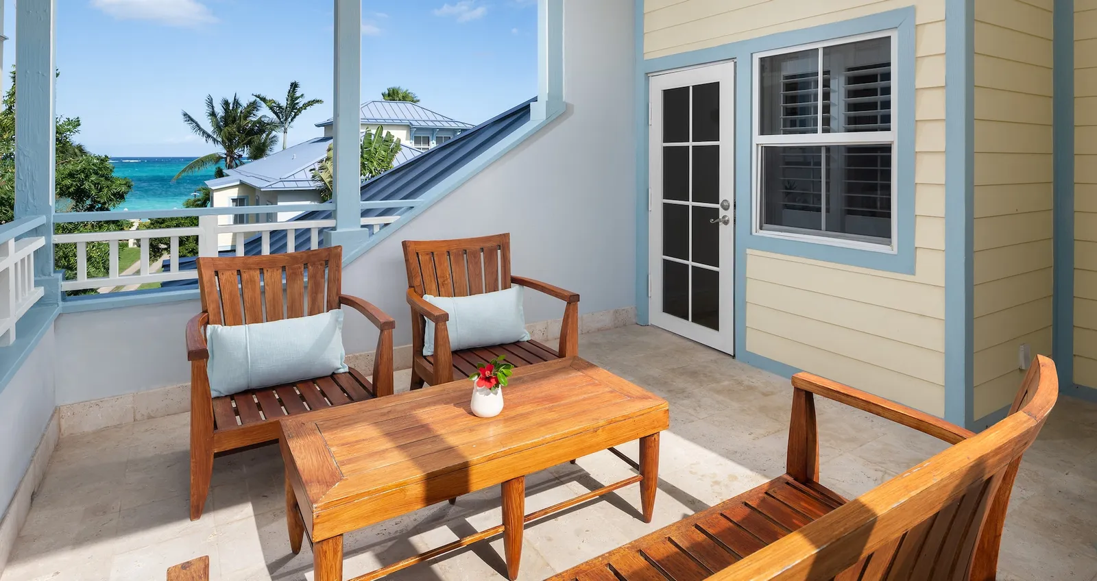 A veranda with teak armchairs and a low table, a yellow clapboard wall and a glimpse of the sea beyond the roofs.